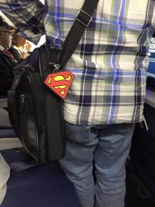 torso of a man in a blue plaid shirt with black computer case over his shoulder with a superman logo hooked on it