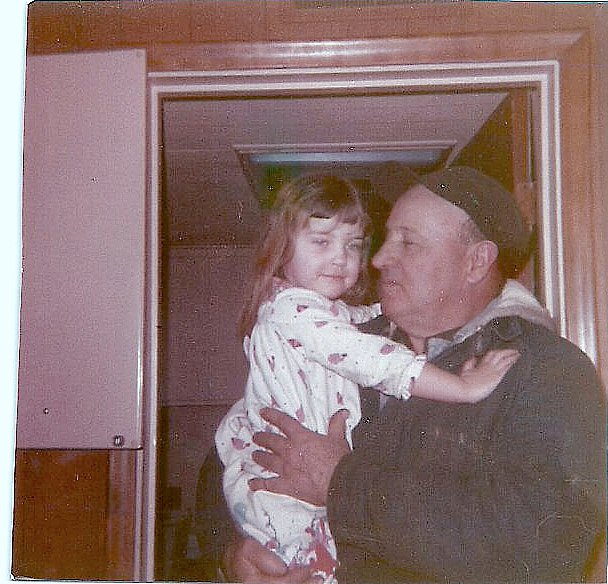 adult male dressed in winter coat and ball cap holding a small girl in pajamas in a kitchen with pink cabinets 